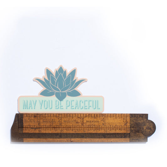 Decorative wooden ruler with a lotus flower and 'May You Be Peaceful' text on a white background