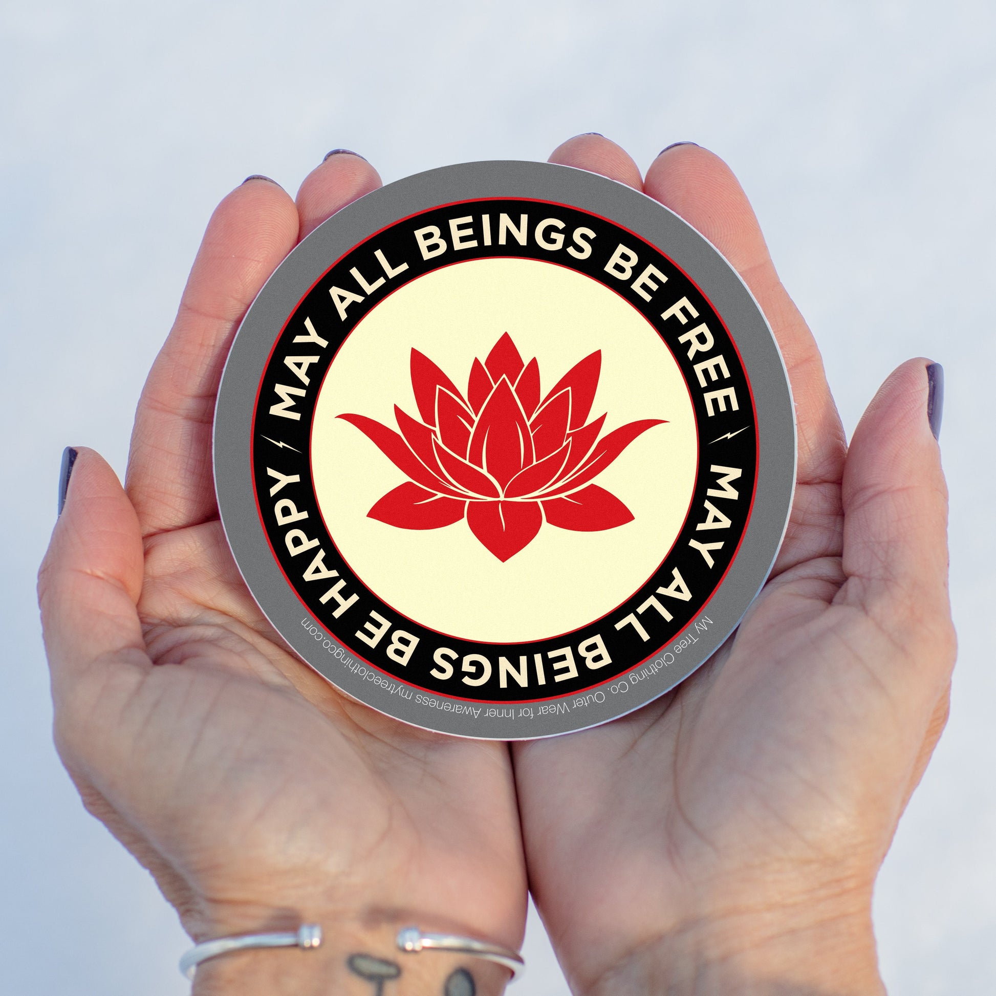Hand holding a circular sticker with a red lotus flower and text may all beings be free may all beings be happy, against a light background.