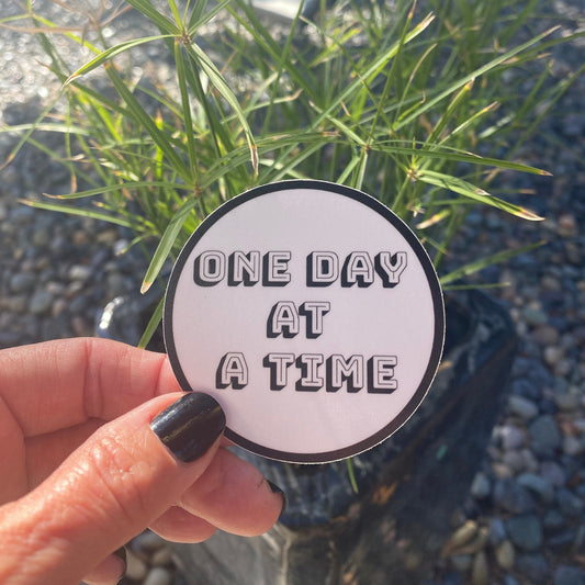 Hand holding a 'One Day at a Time' sticker with greenery in the background