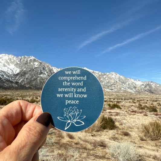 Hand holding a 2.5" blue sticker with a quote from the big book of AA, "we will comprehend the word serenity and we will know peace" and a lotus flower in front of a mountainous landscape.