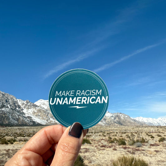 Hand holding a 'Make Racism Unamerican' sticker with a mountainous landscape in the background.