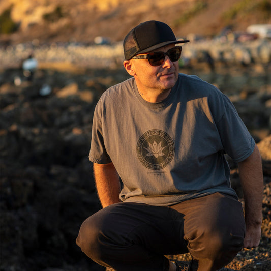 Man wearing sunglasses and a cap sitting on a rocky surface with a blurred background with a gray shirt with a lotus in the center and the text, may all beings be happy may all beings free from the Buddhist loving kindness prayer.