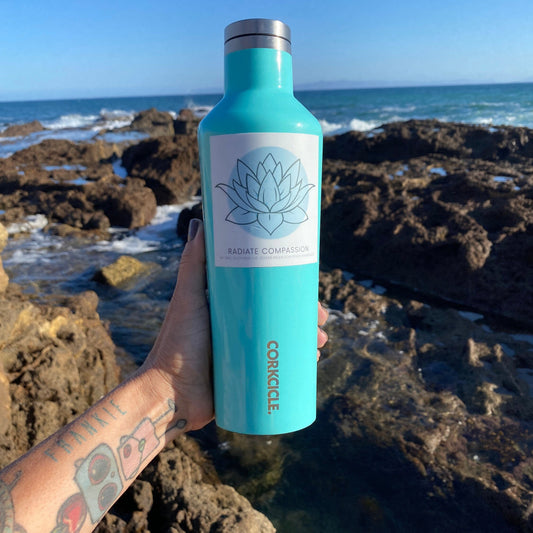 Turquoise water bottle with a 3" square sticker with a blue lotus design and text, radiate compassion, held by a person on a rocky beach.