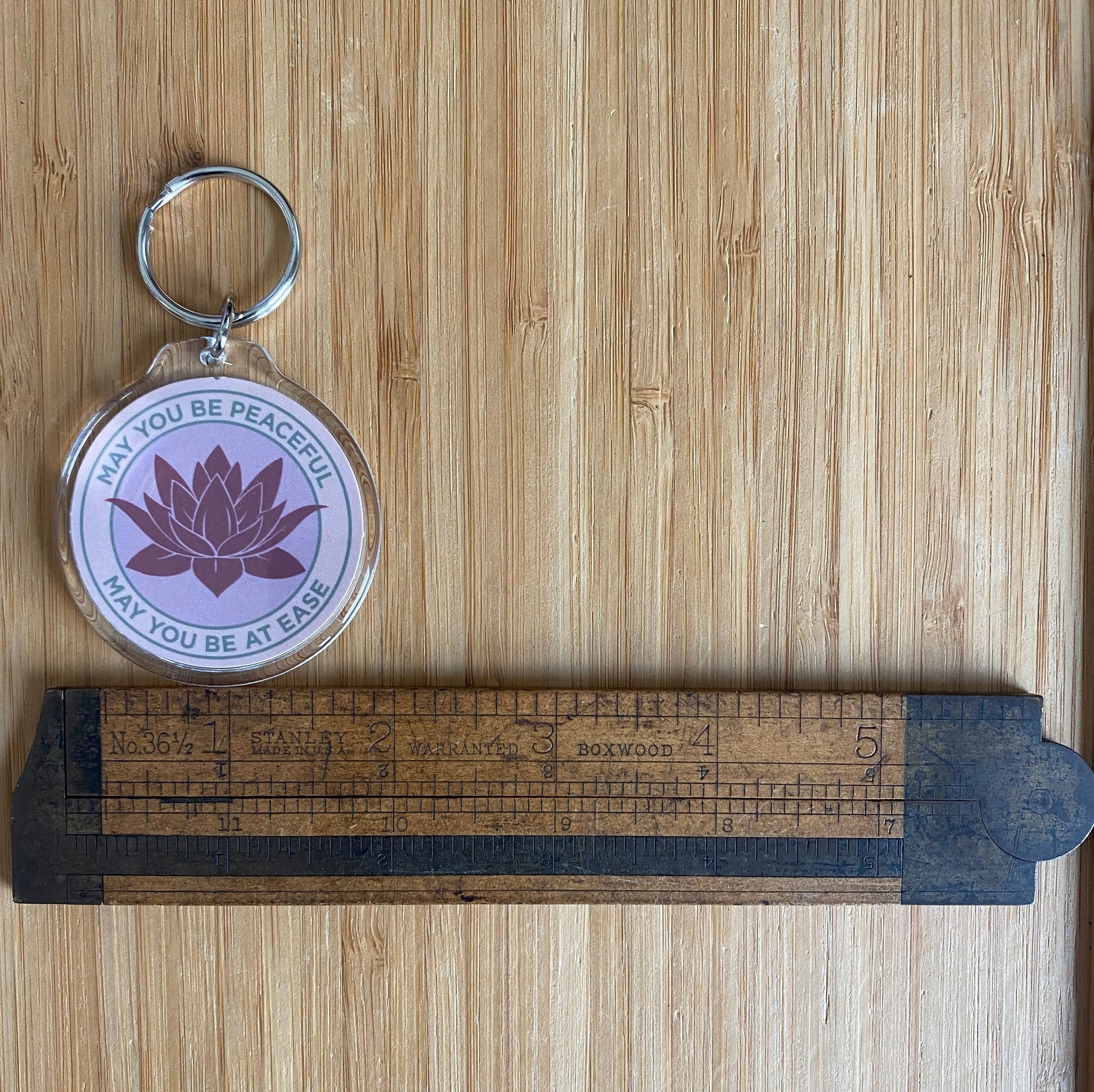 Keychain with lotus flower design and text May You Be Peaceful May You Be At Ease on a wooden surface with a ruler for scale.