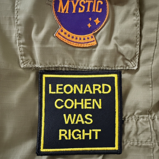Close-up of a beige military-style jacket with two embroidered patches. The lower patch is black with a yellow border and reads 'LEONARD COHEN WAS RIGHT' in yellow text.