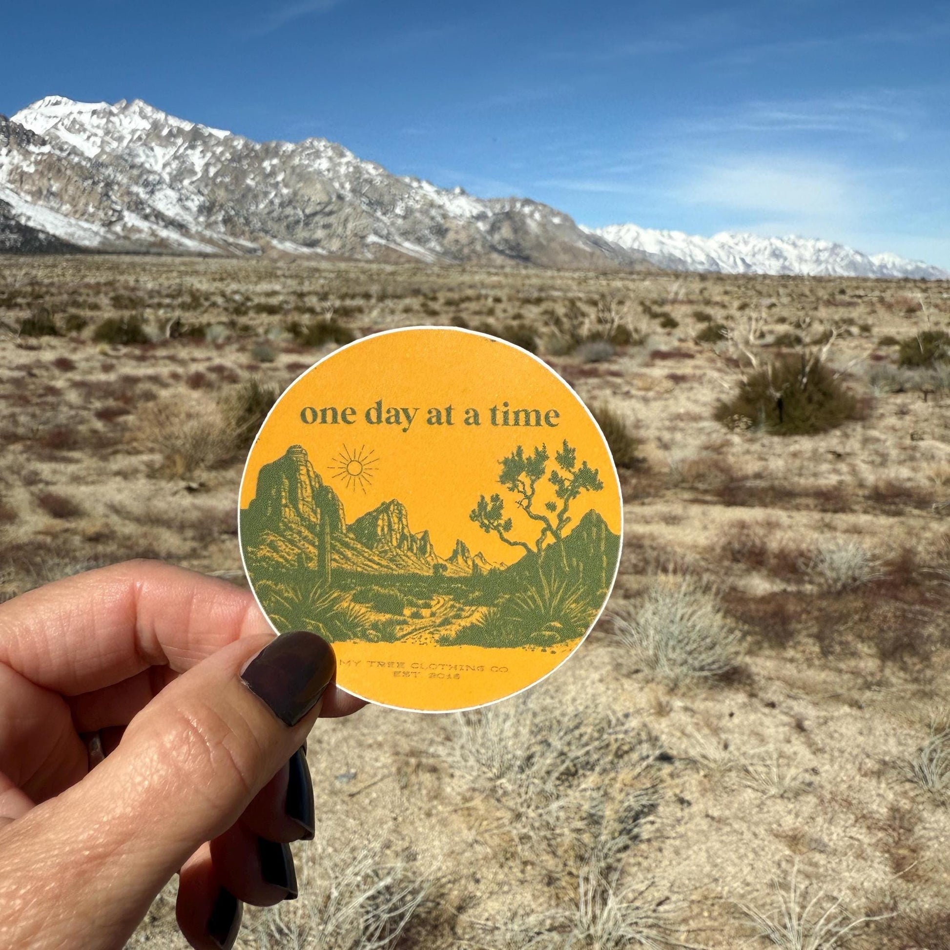 Hand holding a 'one day at a time' sticker with desert landscape design against a mountainous background.