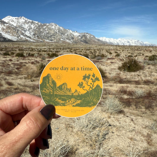 Hand holding a 'one day at a time' sticker with desert landscape design against a mountainous background.