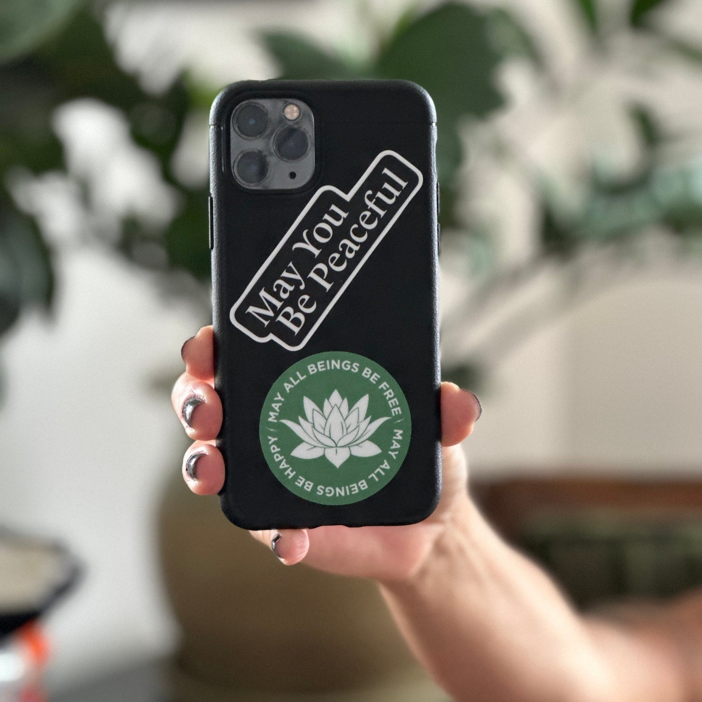 Phone case with 'May You Be Peaceful' text held by a person with a blurred background.