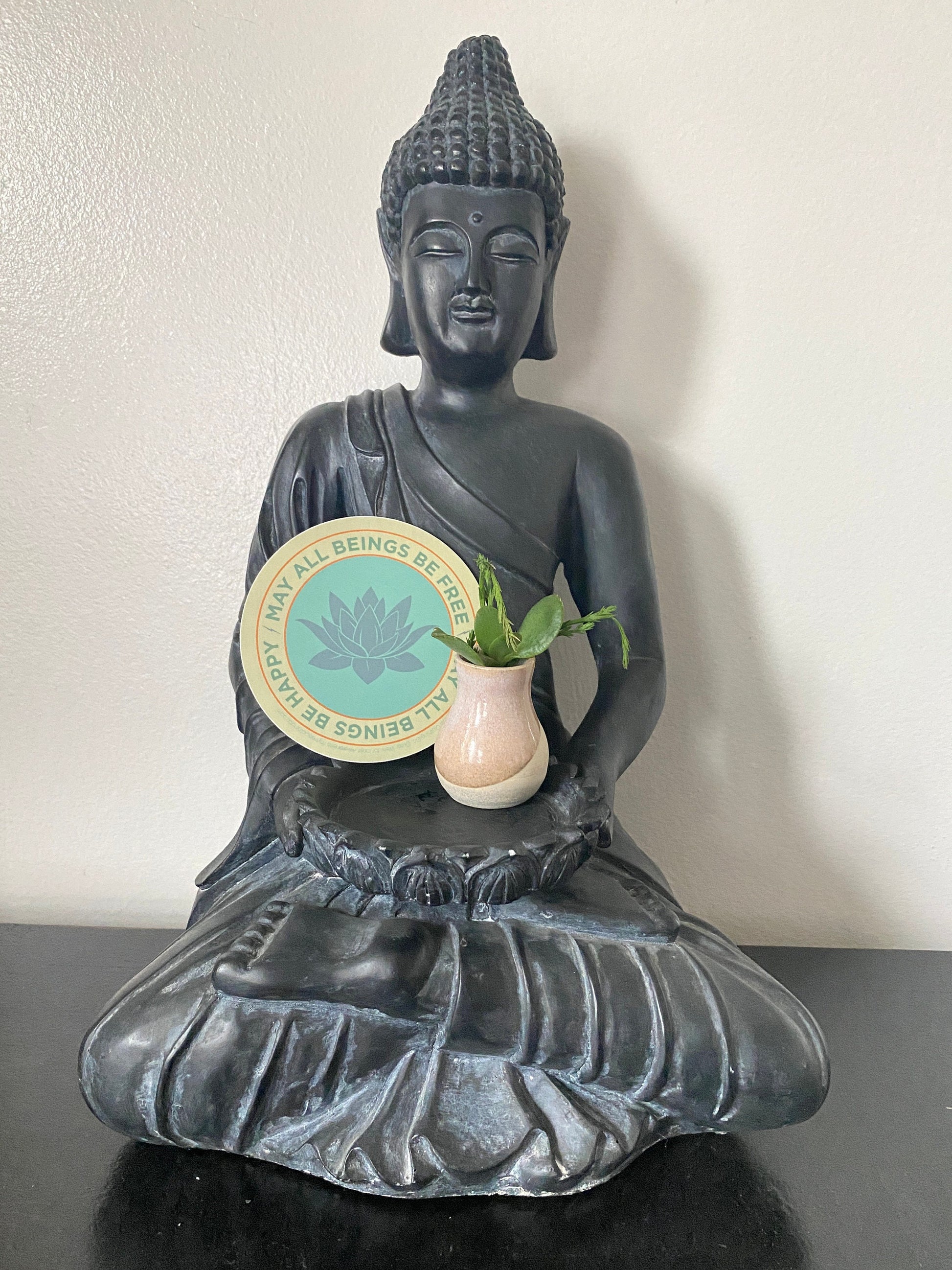 Black Buddha statue holding a vase with a plant on a white background and a 4" round lotus sticker that reads, may all beings be free may all beings be happy.