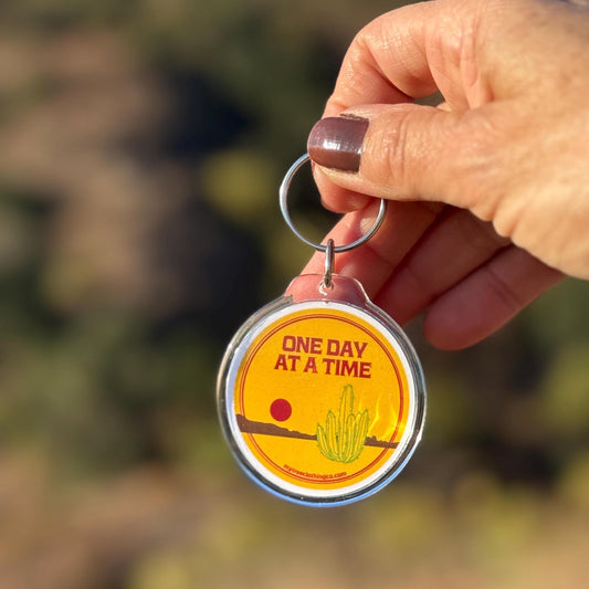Hand holding a keychain with 'One Day at a Time' text and cactus illustration against a blurred natural background.