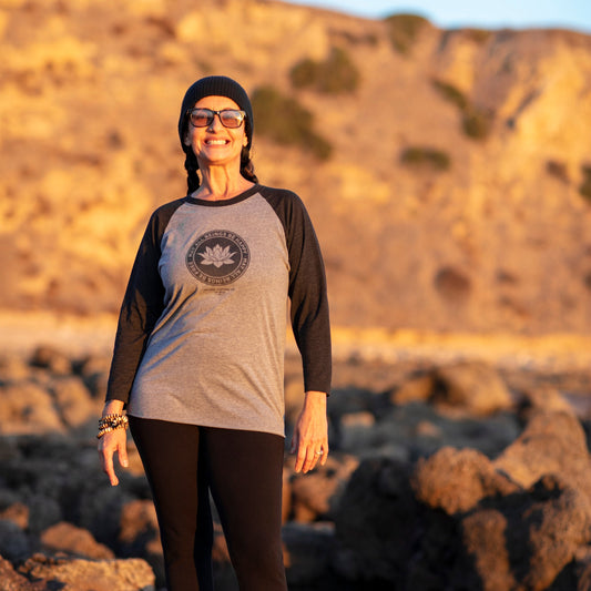 Person wearing a gray and black 1/4 length sleeve shirt with a lotus logo, and the text, may all beings be free may all beings be happy. they're wearing sunglasses, and a beanie standing on rocky terrain at the Pacific Ocean.