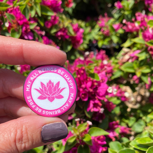 Pink button with lotus flower design, may all beings be happy may all beings be free, held in front of pink flowers
