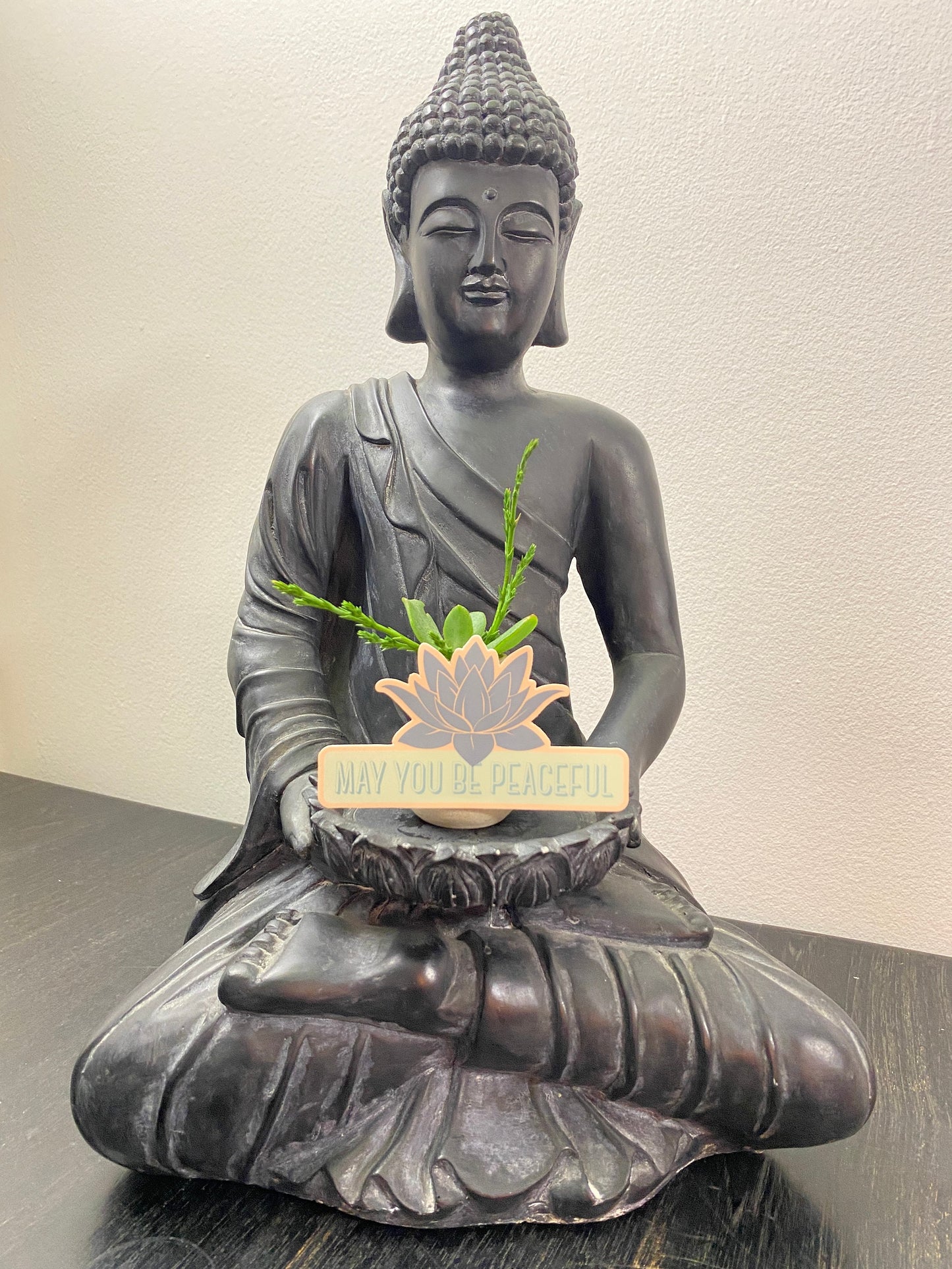Buddha statue with a small plant and a sign on a neutral background with a lotus sticker that reads, may you be peaceful, in teal.