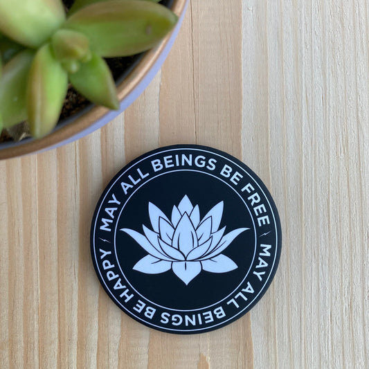 3.5" round black and white magnet with lotus flower design and text, may all beings be free, may all beings be happy, on a wooden surface with a plant.