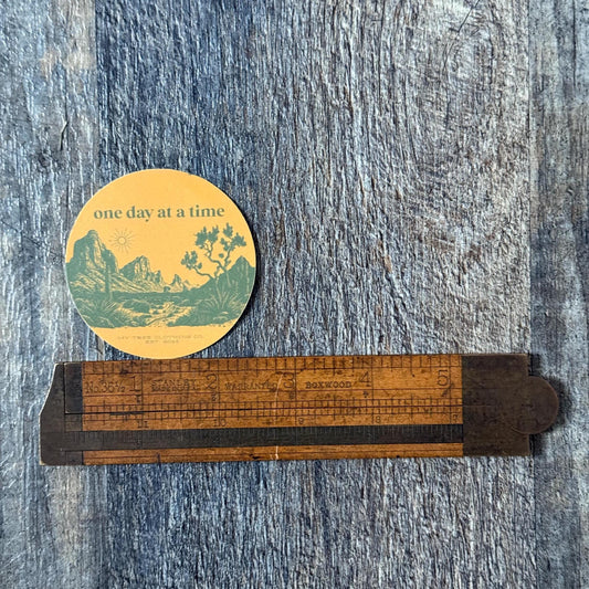 Orange and green 'one day at a time' round sticker with a desert scene and joshua trees on a wooden ruler against a textured blue background.