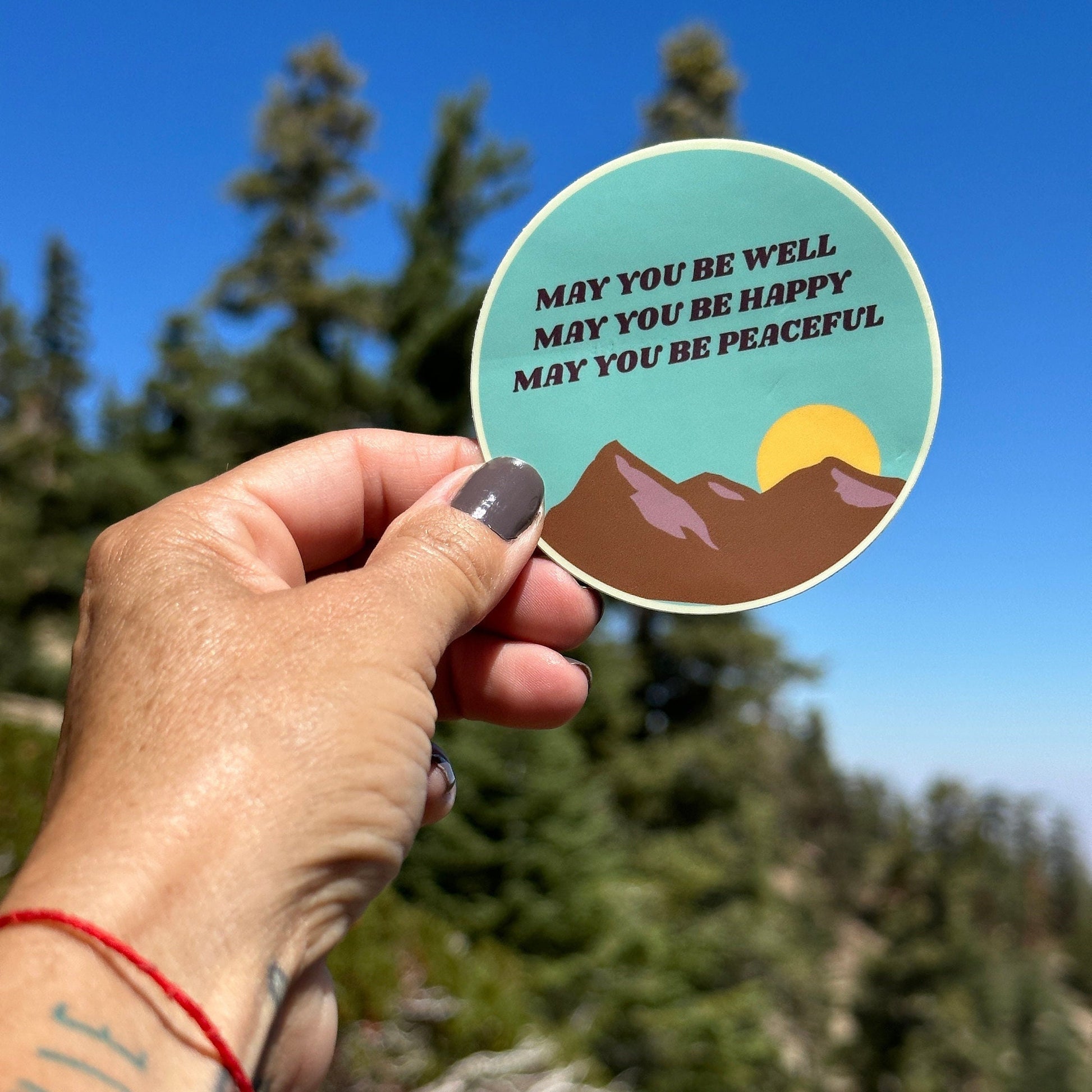 Hand holding a circular sticker with mountain design and Buddhist text, may you be well may you be happy may you be peaceful, against a natural background.