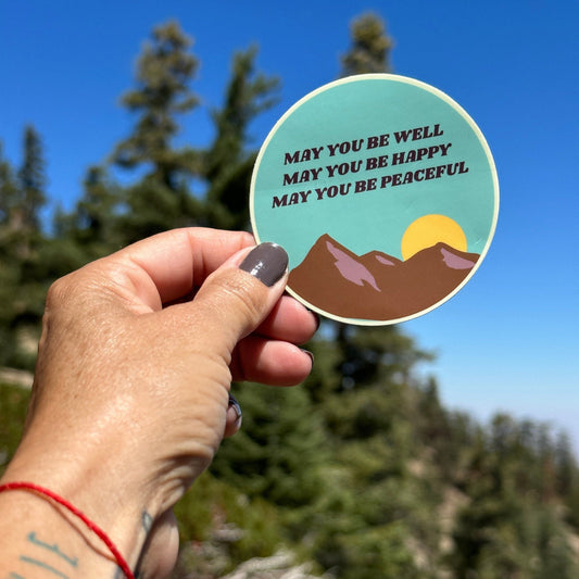 Hand holding a circular sticker with mountain design and Buddhist text, may you be well may you be happy may you be peaceful, against a natural background.