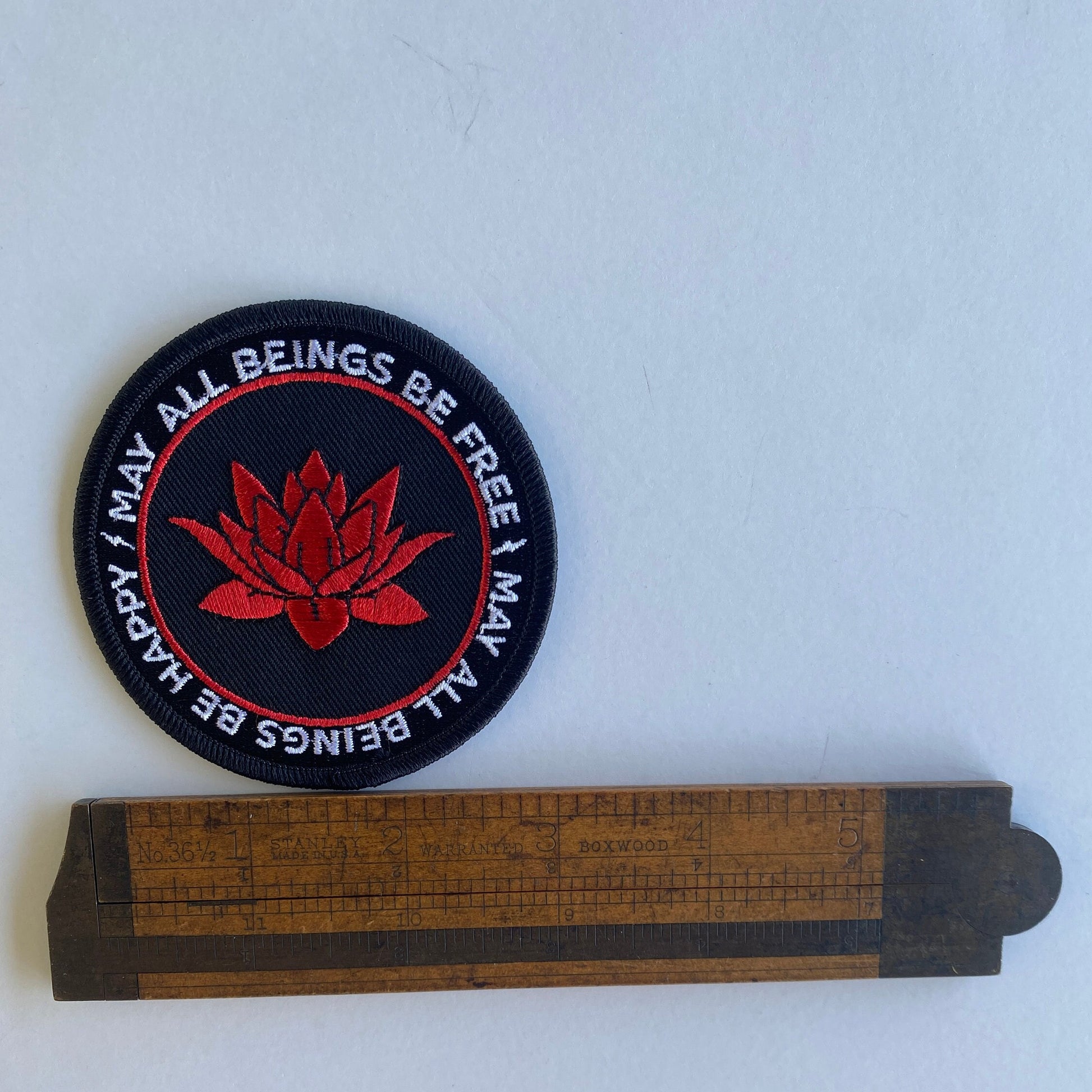 3" round patch with a red lotus flower and the text, may all beings be free may all beings be happy. It is being measured by an antique wooden ruler.