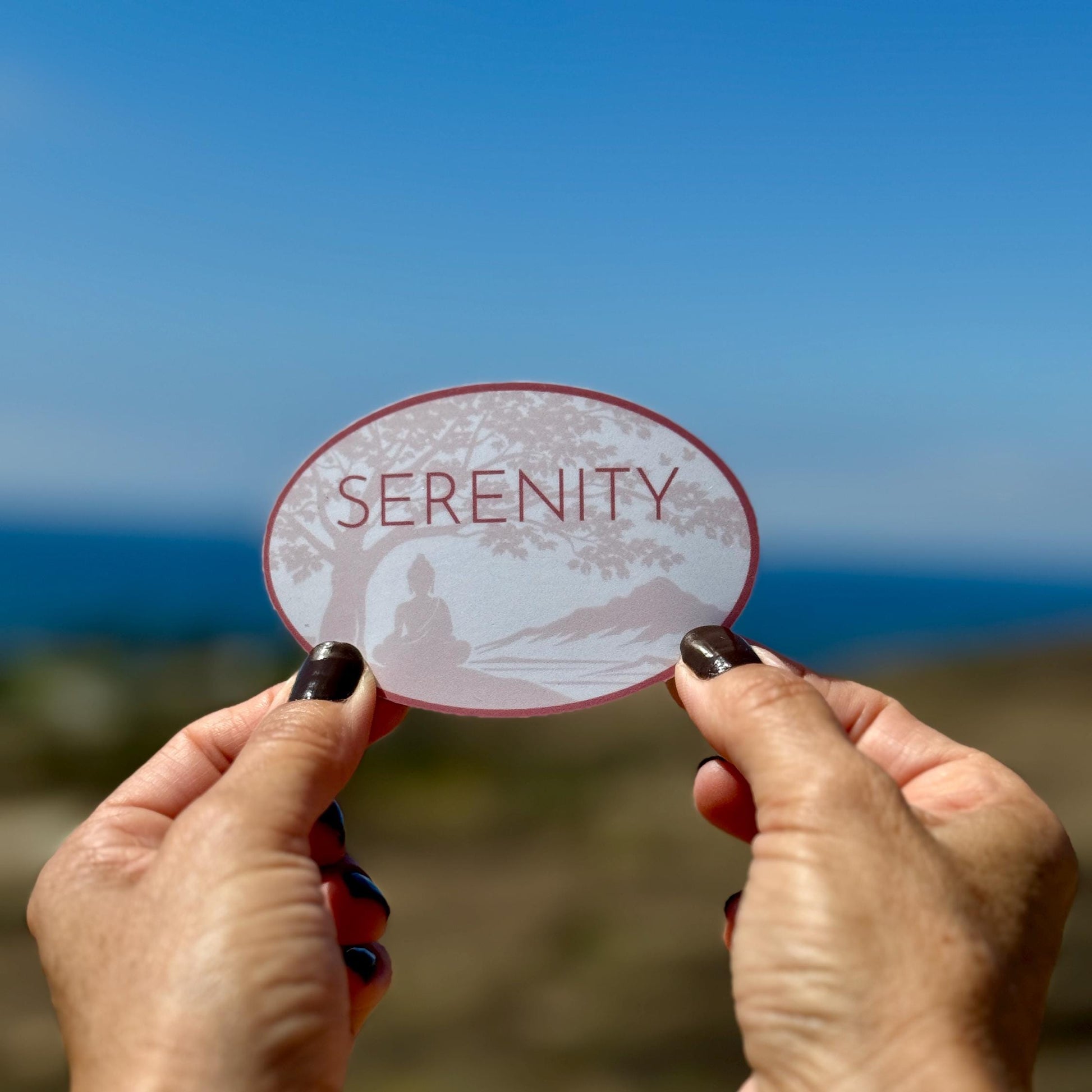 Hands holding an oval shaped 'Serenity' sticker with a Buddha under a Bodhi tree. There is a beach in the background of photo.