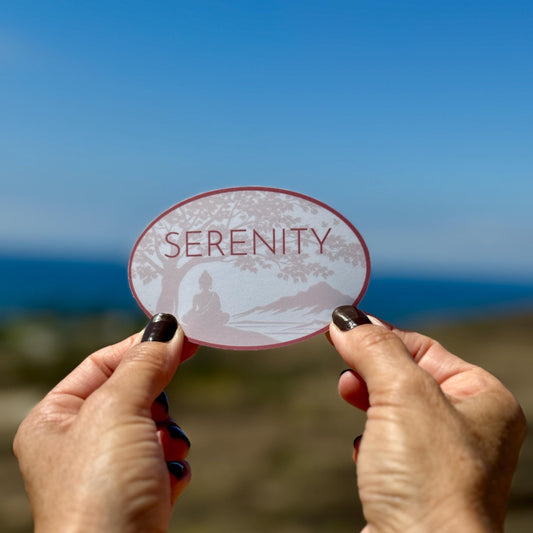 Hands holding an oval shaped 'Serenity' sticker with a Buddha under a Bodhi tree. There is a beach in the background of photo.