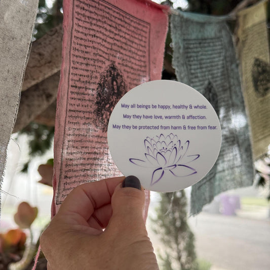 Hand holding a 3" circular sticker with Buddhist prayer and a lotus design in front of prayer flags.