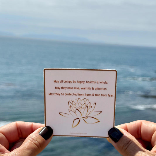 3" square Buddhist sticker with a lotus flower design and text, 'May all beings be happy, healthy and whole. May they have love, warmth and affection. May they be protected from harm and free from fear.' Held by hands against the Pacific Ocean background.