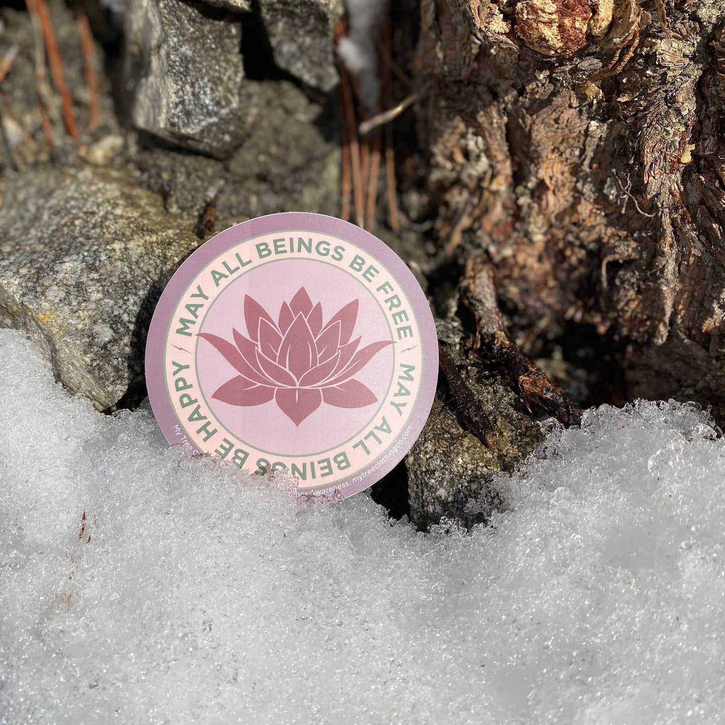 4" Circular sticker with lotus flower design and the text, MAY ALL BEINGS BE FREE MAY ALL BEINGS BE HAPPY on a snowy ground with rocks and pine needles.