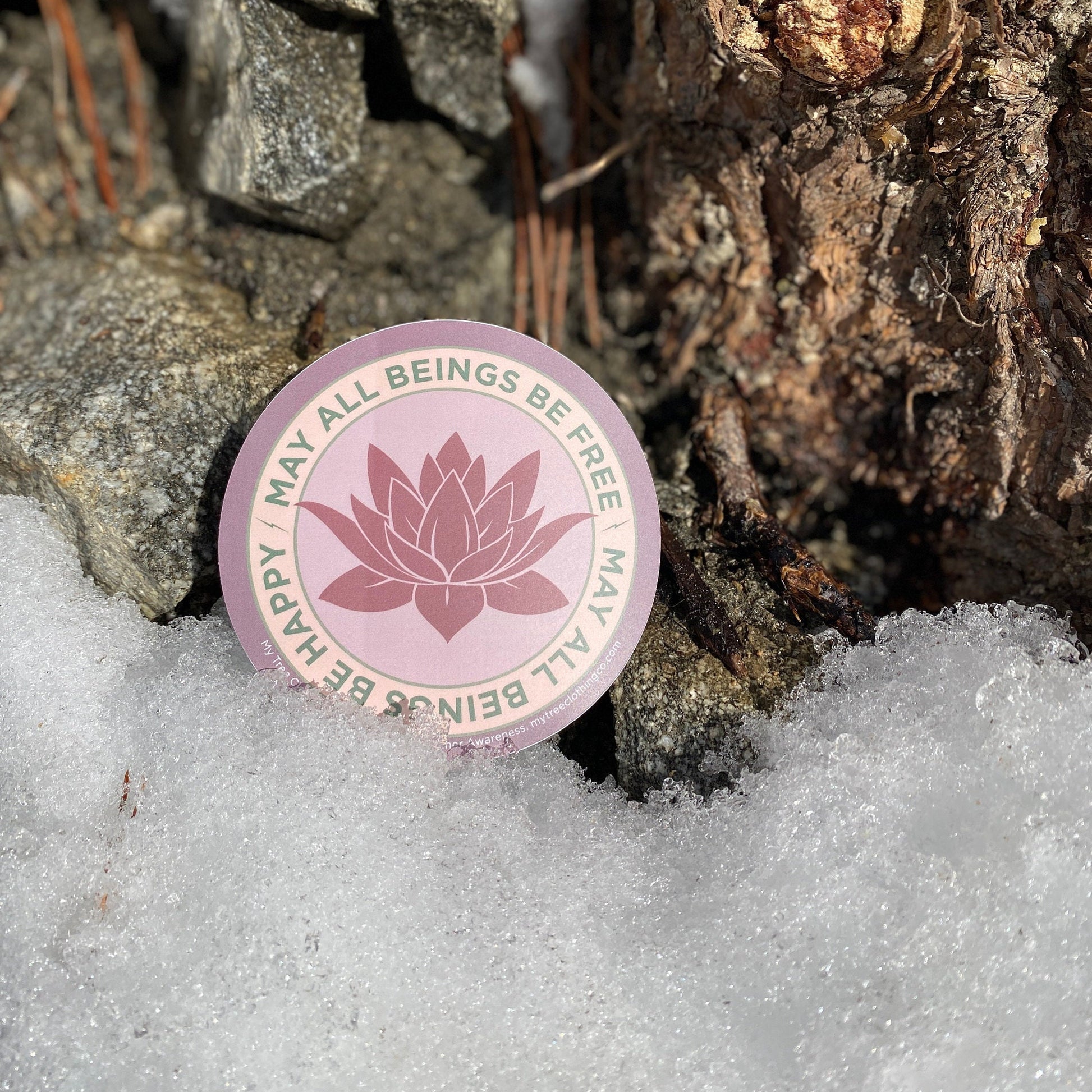 4" Circular sticker with lotus flower design and the text, MAY ALL BEINGS BE FREE MAY ALL BEINGS BE HAPPY on a snowy ground with rocks and pine needles.