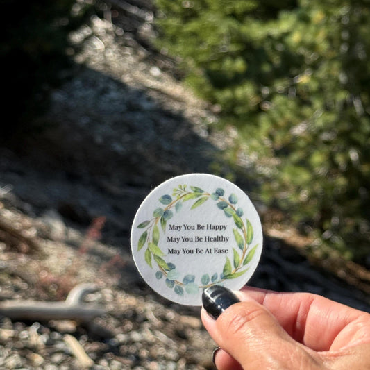 2" round blue sticker with text, may you be happy may you be healthy may you be at ease, held by a hand against a natural background.