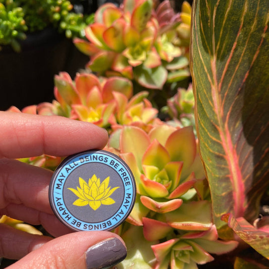 Hand holding a circular badge with a lotus flower design in front of colorful succulents