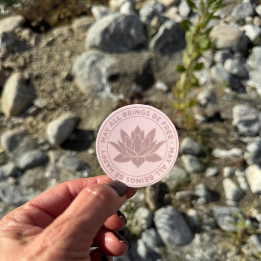 Pink Buddhist lotus flower sticker that reads, may all beings be free may all beings be happy, held by a hand with a natural background of rocks and plants.