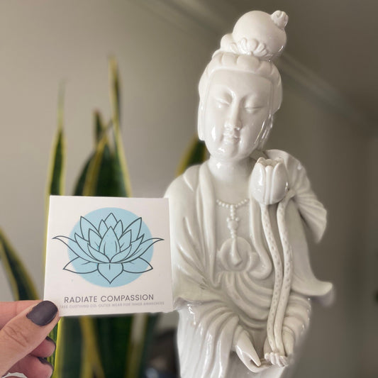 White statue of a Bodhisattva seated figure holding a 3" square sticker with a blue lotus design and text, radiate compassion.