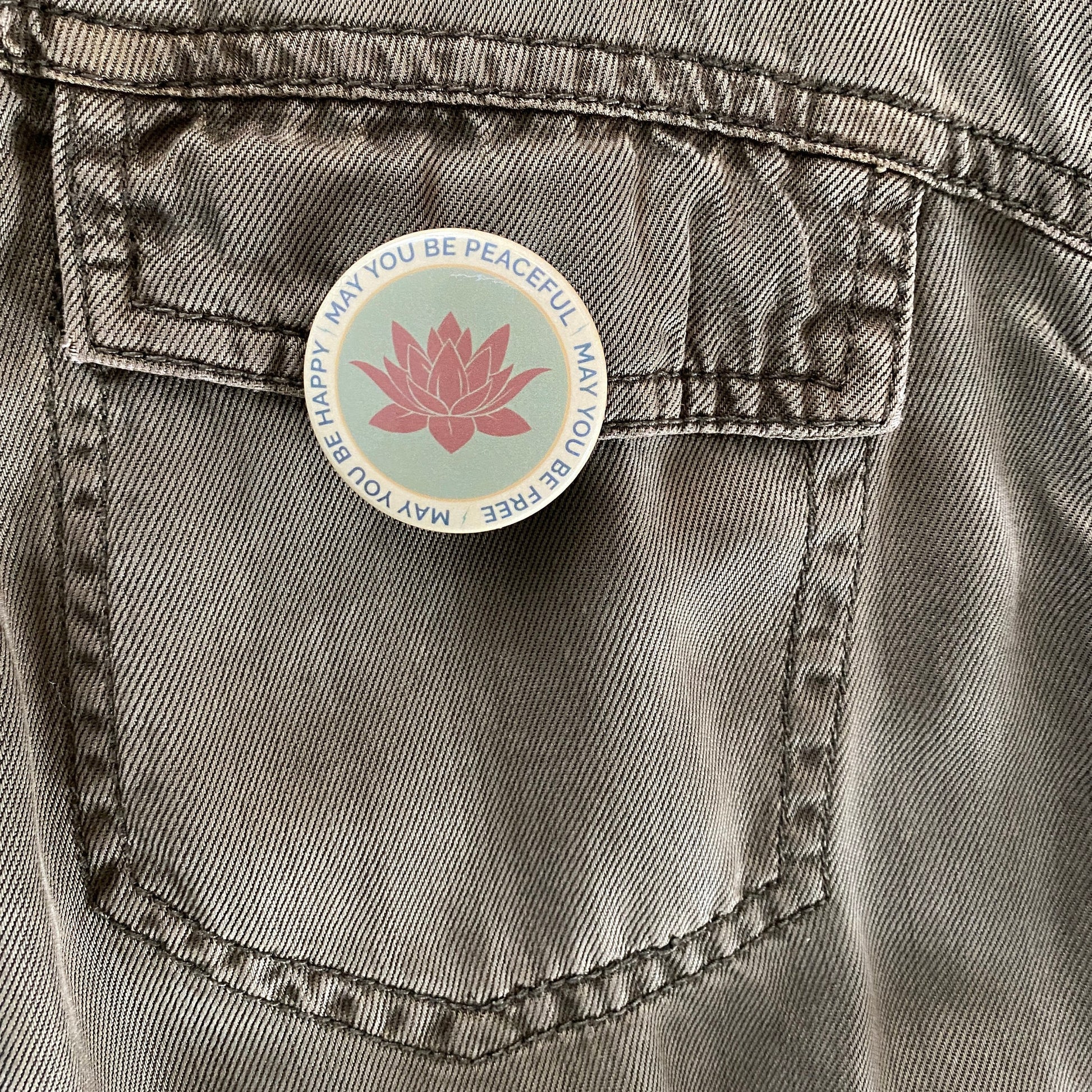 Button with a pink lotus flower design and text, may you be peaceful may you be free may you be happy, on a denim pocket.