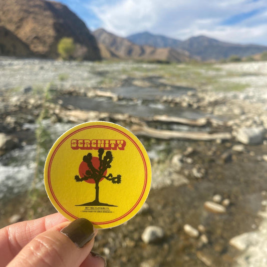 Hand holding a 2" circular sticker with a Joshua tree silhouette, red sun, and 'Serenity' text on a yellow background. against a desert landscape.