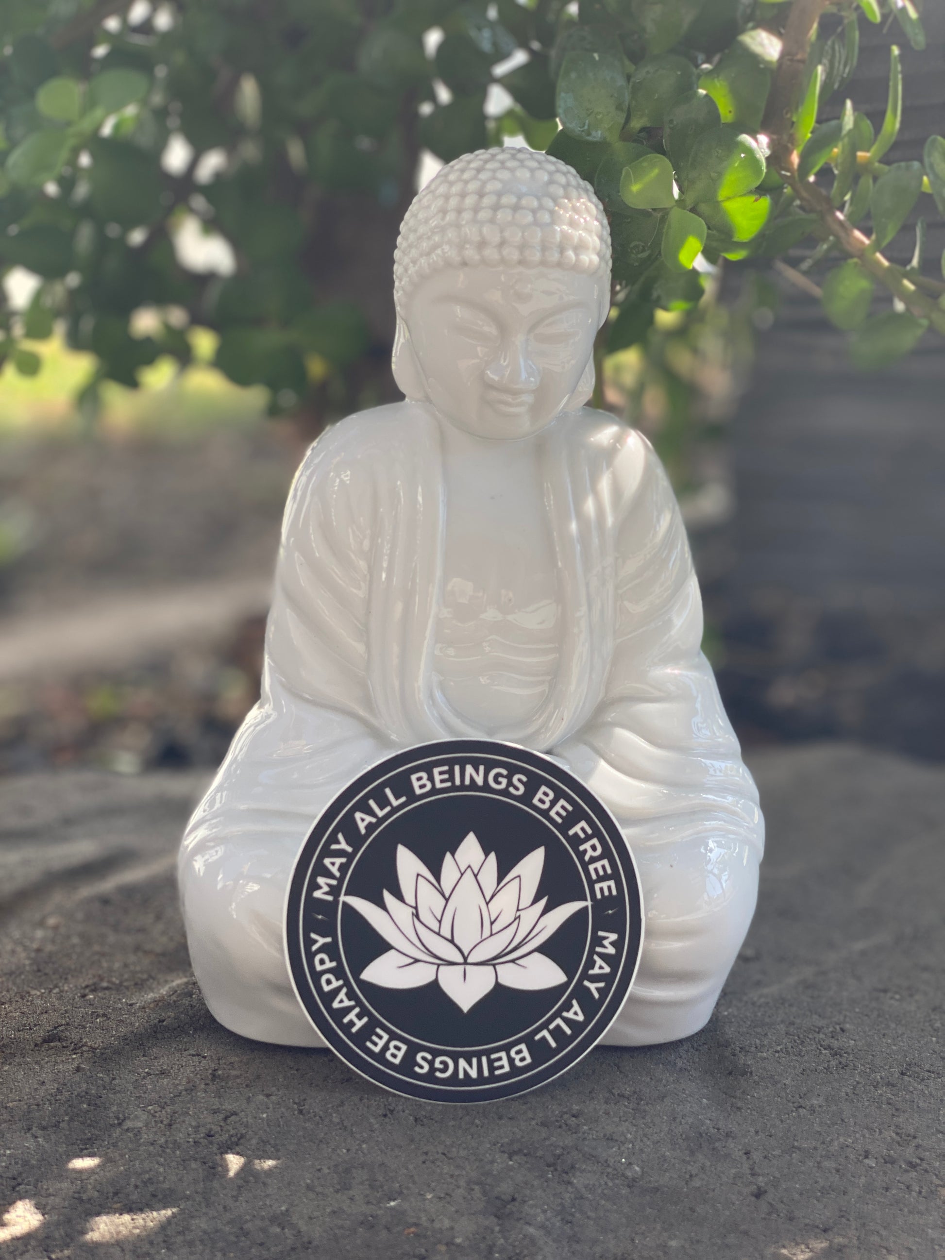 White Buddha statue with a circular emblem featuring a lotus flower and text, may all beings be happy-may all beings be free, on a natural background.