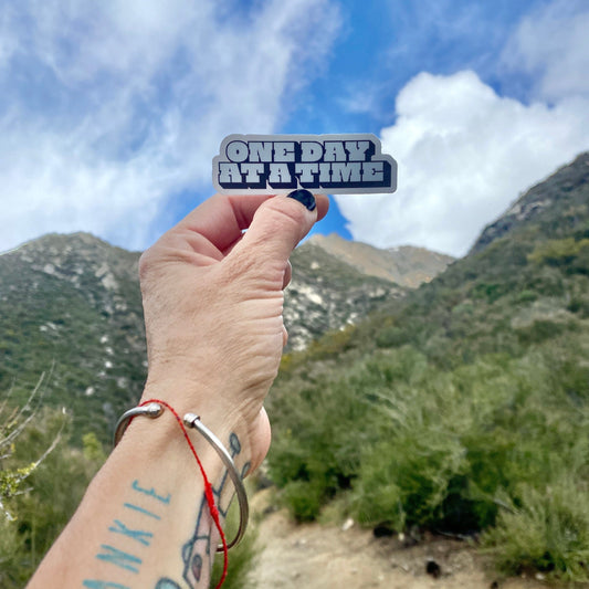 Hand holding a 'ONE DAY AT A TIME' sticker with mountains and blue sky in the background