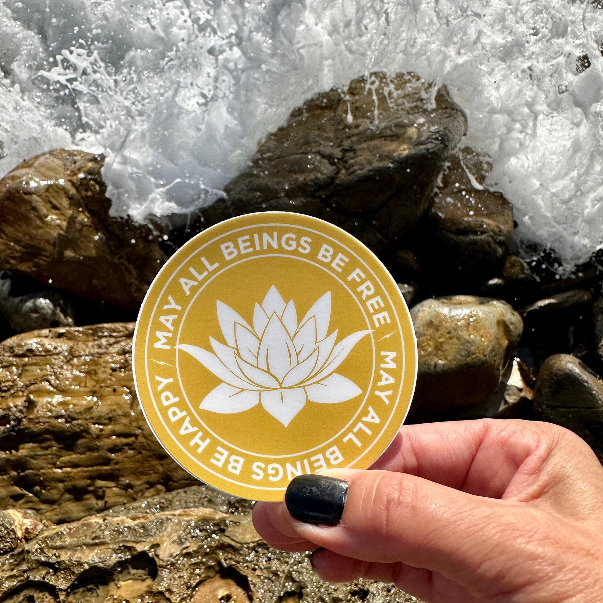 Hand holding a 3" gold and white sticker with a lotus flower design and text, may all beings be free may all beings be happy, against a background of rocks and water.