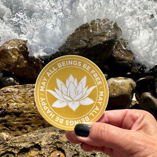 Hand holding a 3" gold and white sticker with a lotus flower design and text, may all beings be free may all beings be happy, against a background of rocks and water.