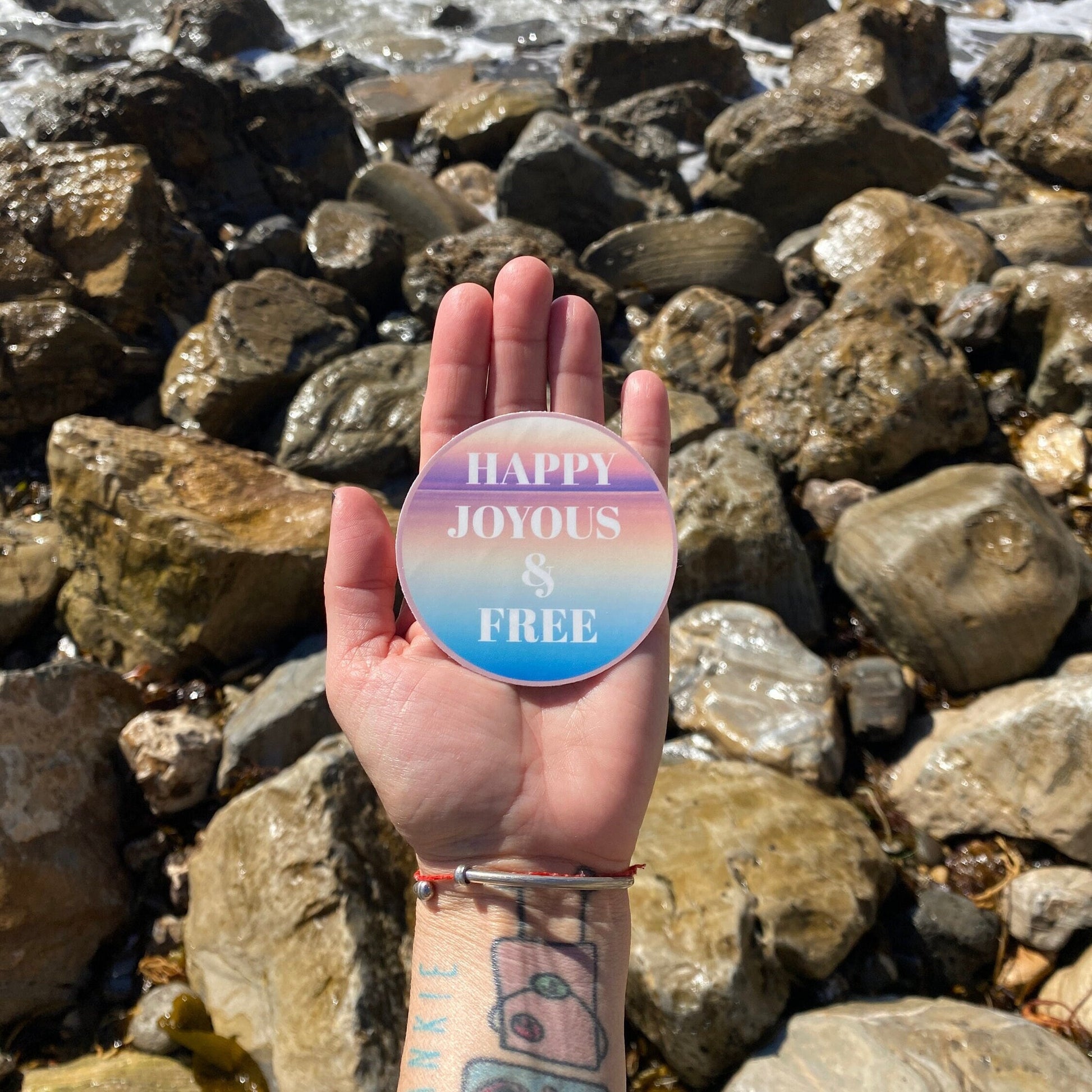 Hand holding a colorful circular sticker with 'Happy Joyous & Free' text against a rocky background.