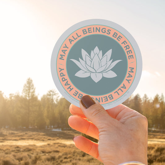 Hand holding a 4" circular sticker with a lotus flower design and text, may all beings be free may all beings be happy, against a blurred natural background.