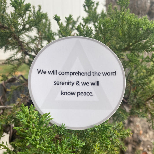 Three inch round white sticker with AA circle and triangle design and the quote, we will comprehend the word serenity and we will know peace. It is in front of a green bonsai tree.