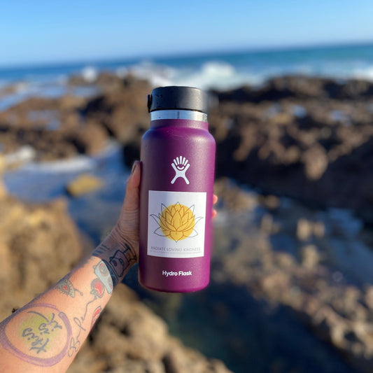 Purple Hydro Flask water bottle with a yellow 'Radiate Loving Kindness' 3" square sticker held by a person against a blurred coastal background.