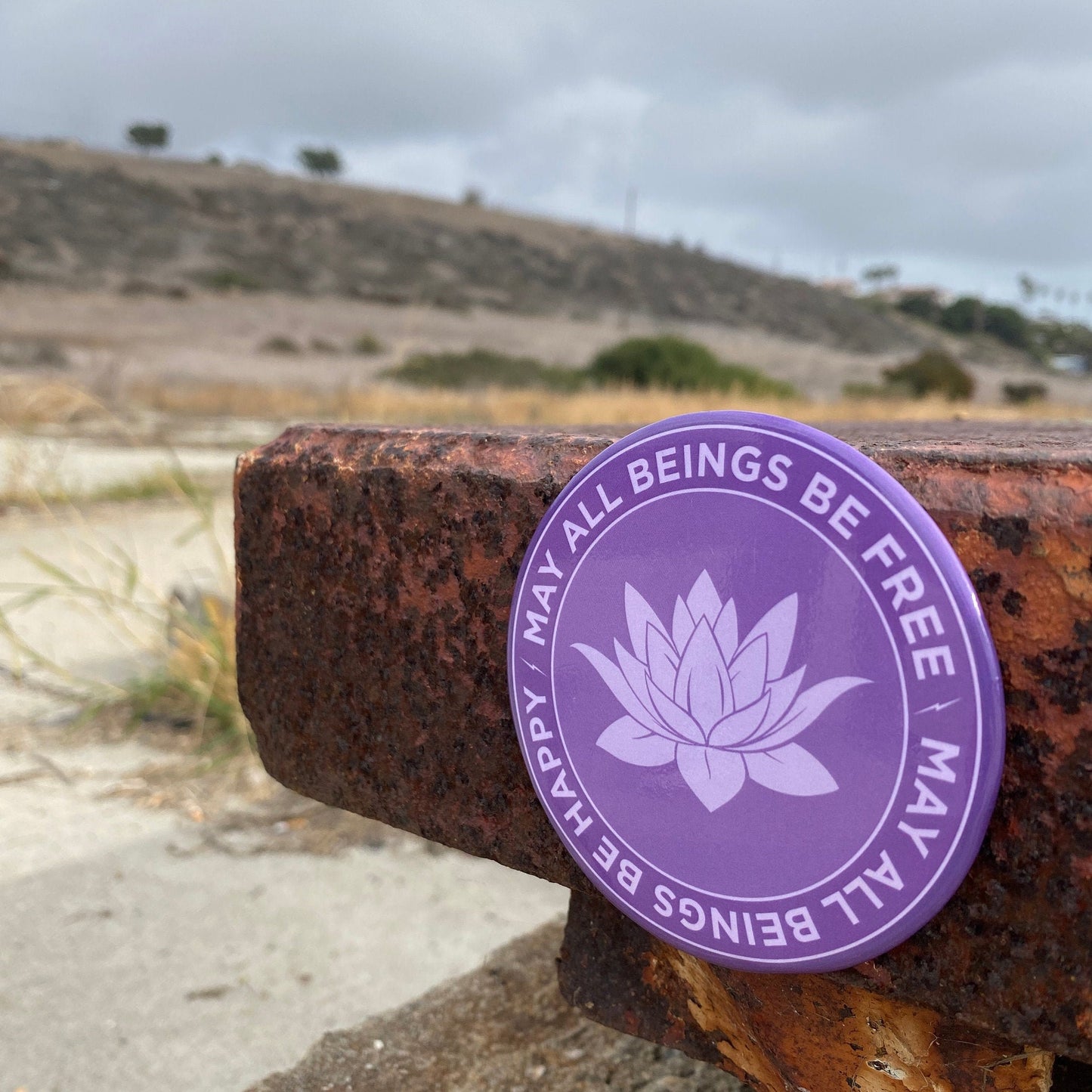 Purple magnet with lotus design and text, may all beings be free may all beings be happy, on a rusty metal surface with a blurred natural background.