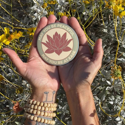 Hand holding a 3.5" lotus flower magnet with the metta prayer, 'may you be peaceful may you be at ease' Surrounded by desert plants.