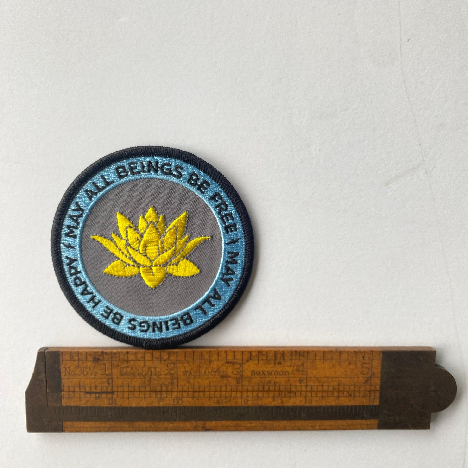 Embroidered patch with a yellow lotus flower and text on a white background, next to a wooden ruler for scale.