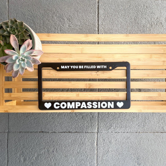 Buddhist prayer black license plate frame with 'May you be filled with compassion' text on a wooden board against a gray wall.