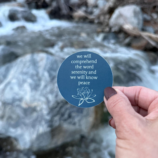 Hand holding a 2.5" blue sticker with a quote from the big book of AA, "we will comprehend the word serenity and we will know peace" and a lotus flower against a natural background