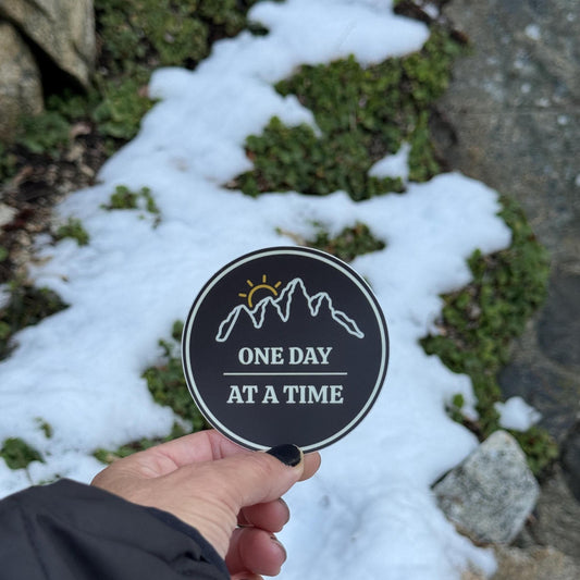 Hand holding a "3 round sticker with outline of mountains and a sunset with 'One Day at a Time' text against a snowy ground background.