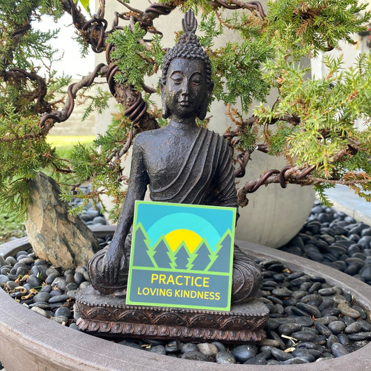 Buddha statue with a square sticker with trees and a sunrise that reads, 'Practice Loving Kindness' in a garden setting.