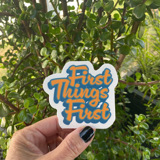 Hand holding a 12 step recovery slogan 'First Things First' sticker with green foliage in the background.